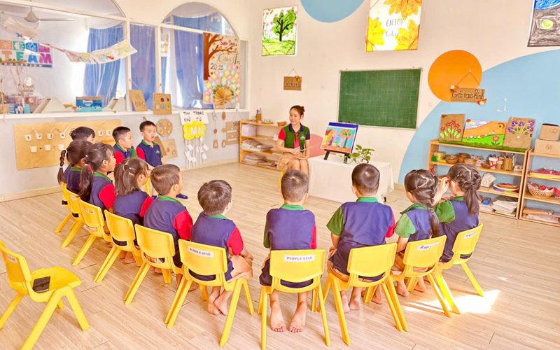 Cả lớp ngồi ghế vàng thành vòng tròn nghe cô trong phòng học sáng màu